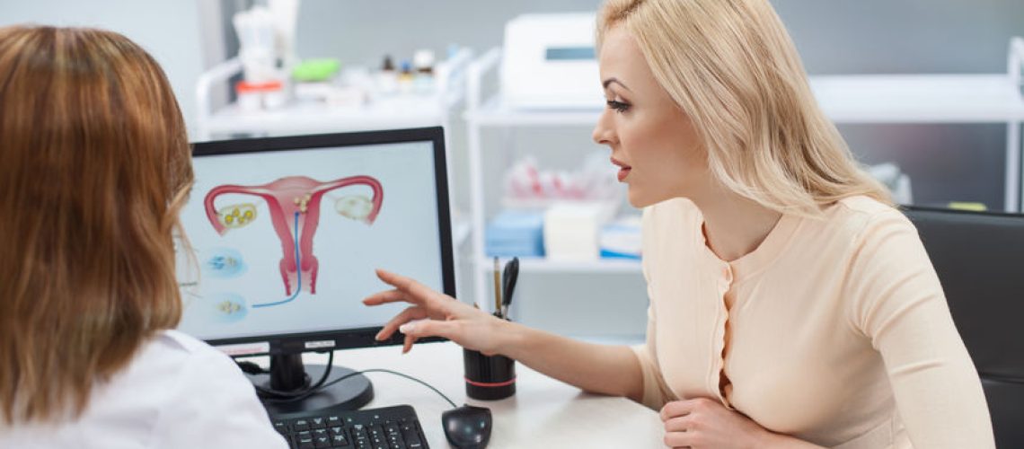 Cheerful young woman is asking advice in her gynecologist. She is sitting at the desk and pointing finger at the computer with uterus picture. Lady is listening to doctor with concentration
