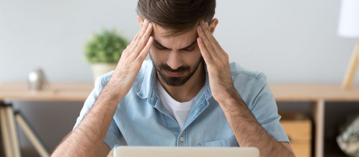 Tired man suffering from headache after long work with laptop, massaging temples, exhausted freelancer or student feeling pain or blood pressure, unwell, health problem concept, taking break